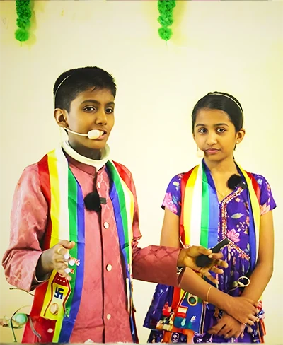 Jainam and Jivika presenting on stage with headset microphones.